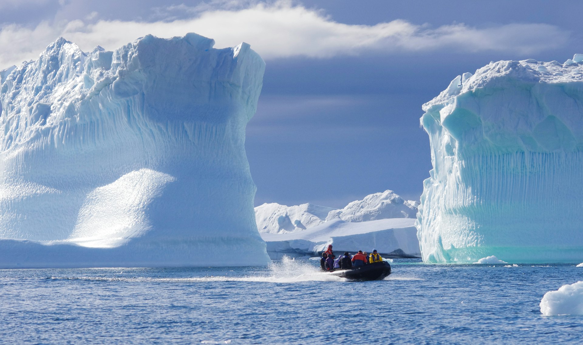 A Zodiac of expedition guests motors past towering blue icebergs in open Antarctic water under a bright sky